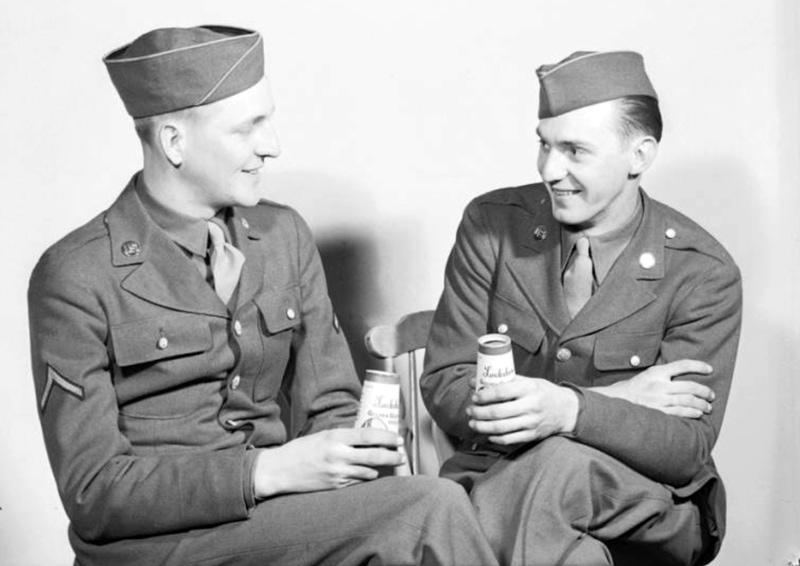 Two soldiers in uniform and caps, seated, drinking Lockshore brand milk from half-pint paper milk containers manufactured by Sutherland Paper Company.