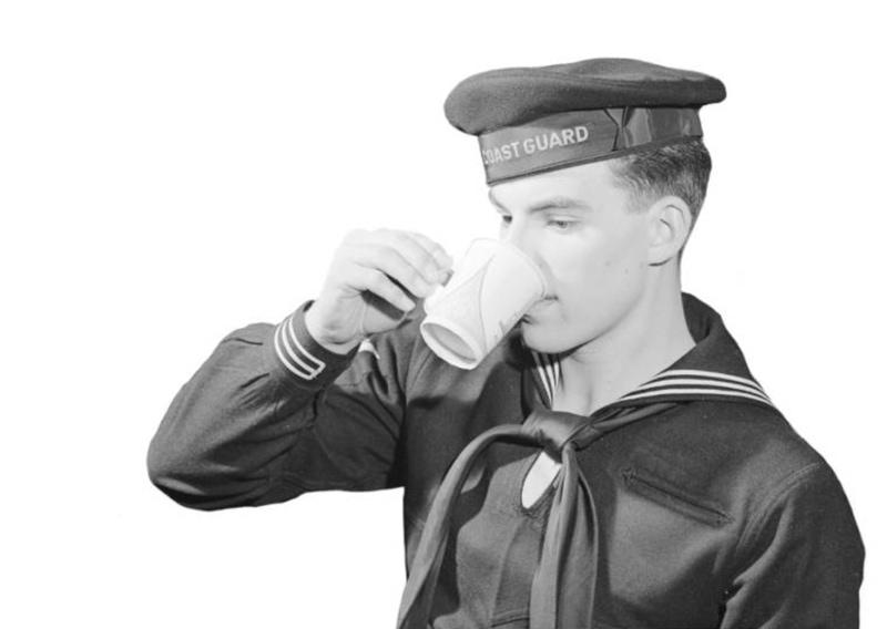 Member of U.S. Coast Guard in uniform and cap, drinking from paper cup with handle manufactured by Sutherland Paper Company.